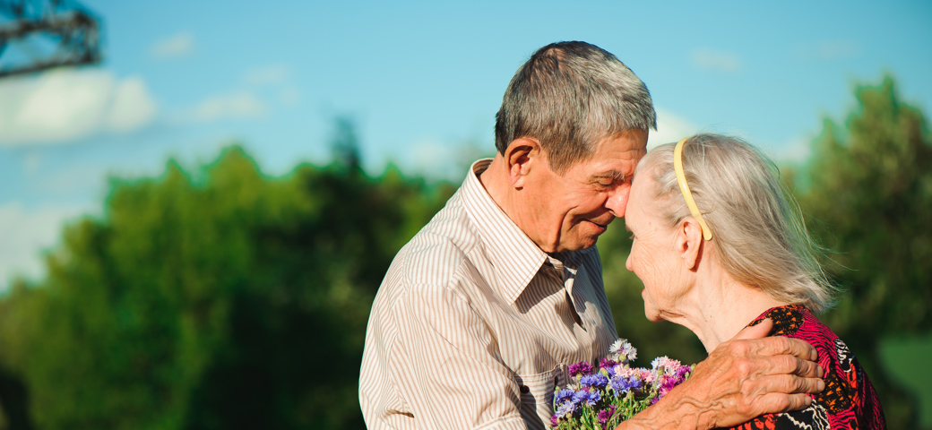 Senior mand og kvinde står holder om hinanden, mens kvinden holder blomster imellem dem