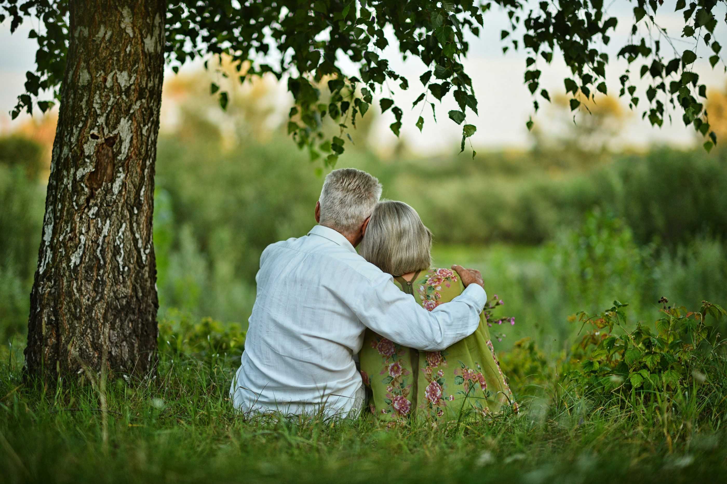 Seniorpar, der sidder under et træ og holder om hinanden mens de kigger væk fra kameraet på et grønt område